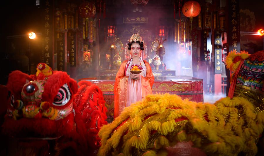 Mujer con vestimenta tradicional china y tocado adornado sosteniendo un cuenco con naranjas dentro de un templo místico, acompañada por dos coloridos trajes de la danza del león en primer plano.